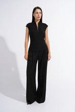 Load image into Gallery viewer, A woman modeling a black sleeveless top with a split neckline and matching black wide-leg trousers, standing against a plain white studio background.
